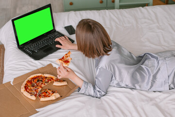 Young brown-haired woman lies in gray pajamas in bed in the bedroom On isolation in the house Looks at a laptop and eats pizza next to a mobile phone