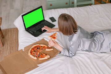 Woman in pajamas in bed Isolation in apartment looking at laptop holding pizza in hand