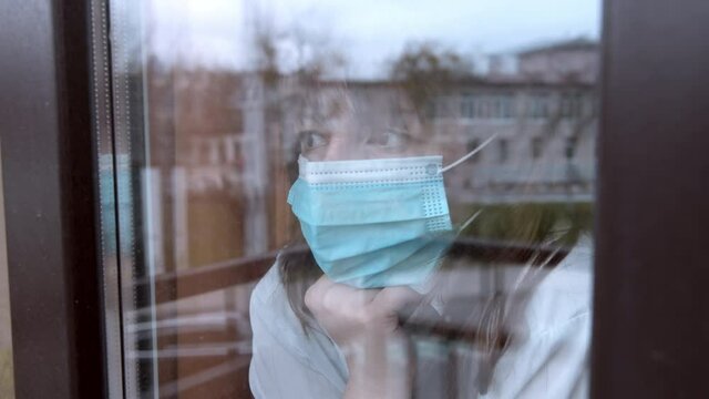 sad woman in a mask looks out the window assesses the situation, takes off the medical mask, the end of the pandemic, mask fatigue, cinematic shot