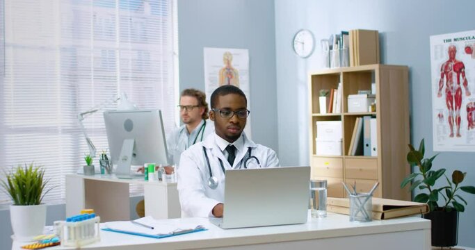 Young African-American doctor with glasses sits in hospital office and works intently on laptop studying patients' appeals online.