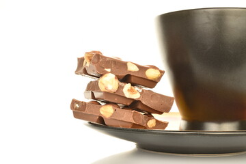 Several slices of dark chocolate with nuts on a saucer with a ceramic black cup, close-up, isolated on white.