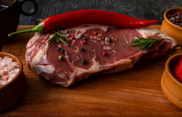 Raw piece of ribeye beef meat on wooden cutting board on a wooden table, with hot peppers, peppercorns mixture, paprika, himalayan salt. Fresh food. Everything for a barbecue. Macro