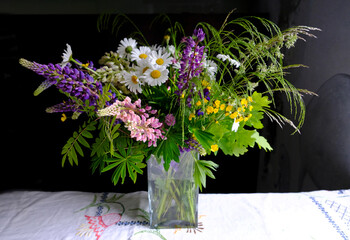 Bouquet of summer solstice flowers and greens in a square glass vase on a wooden table