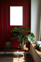 A monstera flower stands in a wicker basket on a coffee table against a red wall. A white frame hangs on the wall. A place for a picture or poster. Modern interior. Sunlight shines on the wall.