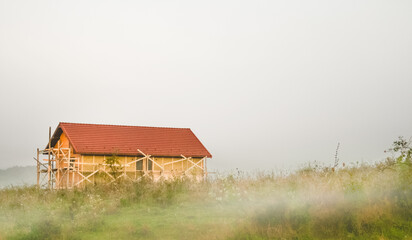 Morning landscape with construction of new wooden cottege house for tourists in the Carpathian mountains, chalet style building with the foreground of meadow covered by fog.