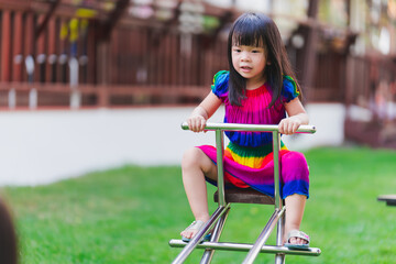 Asian child in colorful dress playing seesaw in the laws playground at outdoors. Cute girl is happy...