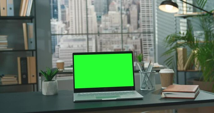 Full length view of the laptop with green blank mockup screen is on top of wood table with modern office at the background. Technologies concept