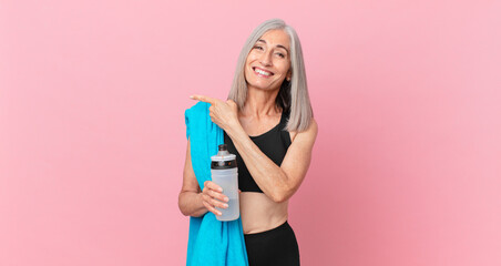 middle age white hair woman smiling cheerfully, feeling happy and pointing to the side with a towel and water bottle. fitness concept