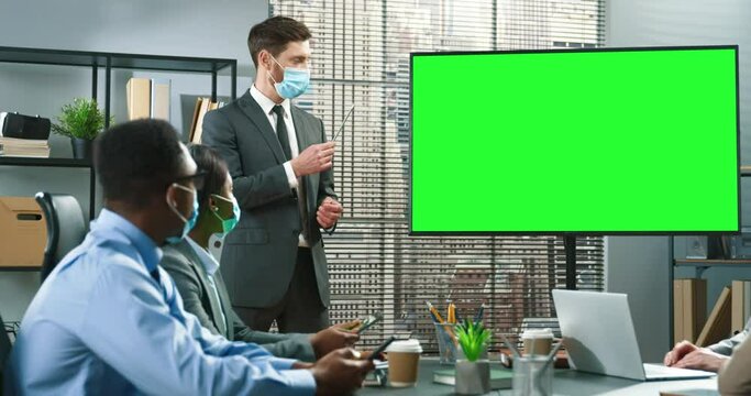 Full length view of colleagues are working together in office. Caucasian man wearing protective mask is standing near the projector and telling to his colleagues. Man feeling confidently while saying