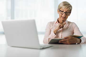 Mature businesswoman working on laptop and taking notes in the office