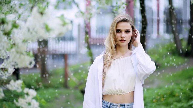Portrait of young beautiful blonde woman in the flowered garden in the spring time. Attractive fashion model walking in park. Blossoms fall from trees. Slow motion
