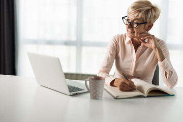 Smiling mature businesswoman working on laptop and taking notes in the office