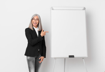 middle age white hair woman looking excited and surprised pointing to the side and a board copy space. business concept