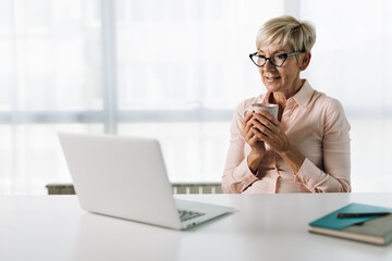 Mature businesswoman drinking coffee while surfing the Internet on a computer in the office