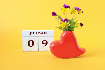 Calendar for June 9 : the name of the month of June in English, the numbers 0 and 9, a vase in the shape of a red heart with a bouquet of violets on a yellow background