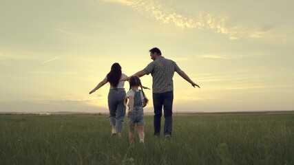 A happy family runs across the field with a small child at sunset in the sky, running mom, dad and kid are jumping cheerfully in the field in the evening, the team is traveling cheerfully playing