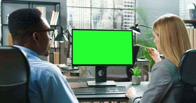 Two multiracial workers sitting at office and discussing business issues together. Blonde woman pen and typing at the computer green screen at the workplace. People looking at each other