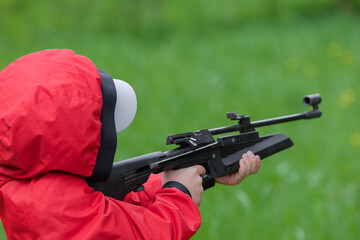 A boy in a red windbreaker and a cap is holding an air gun. The concept of weapons and children,...
