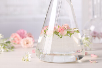 Flask with rose flowers on white wooden table, closeup. Essential oil extraction