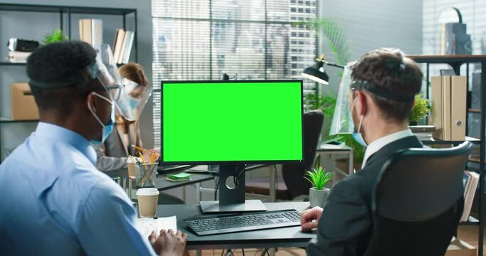Two business people wearing protective masks sitting together at the table in the modern office, talking and looking at the green screen of the computer. Caucasian man explaining something to his