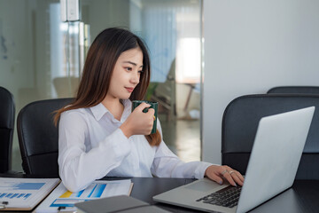 Obraz premium Young Asian businesswoman sitting in the office drinking coffee and working using a laptop and graph.