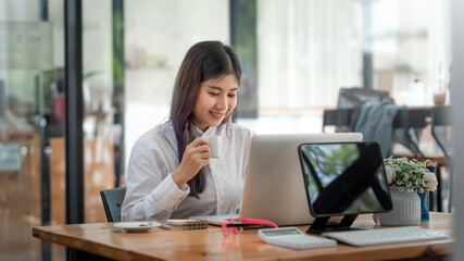 Smiling young Asian woman working on laptop and drinking coffee at office.