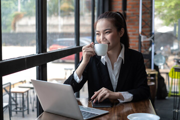 Happy Asian businesswoman sitting and drinking coffee the laptop is placed on the café table looking out the window.