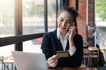 Obraz premium Front view of a beautiful businesswoman holding a credit card while talking on the phone to order online using laptop placed on the office desk.