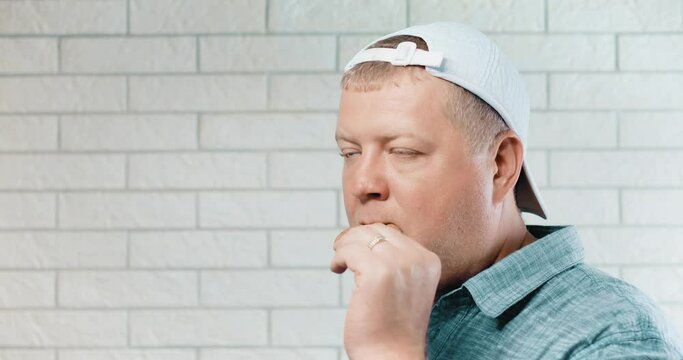 A man stands against a white brick wall and nonchalantly eats a round cookie. The man is wearing a white baseball cap backwards on his head. He is calm and just bites and chews cookies.