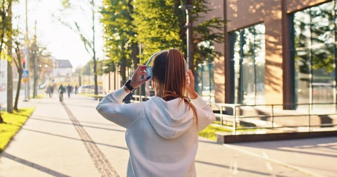 Back view of young female walking outdoors on street putting on headphones and listening to music in good mood. Caucasian woman having walk in city on sunny day enjoying song, rear concept