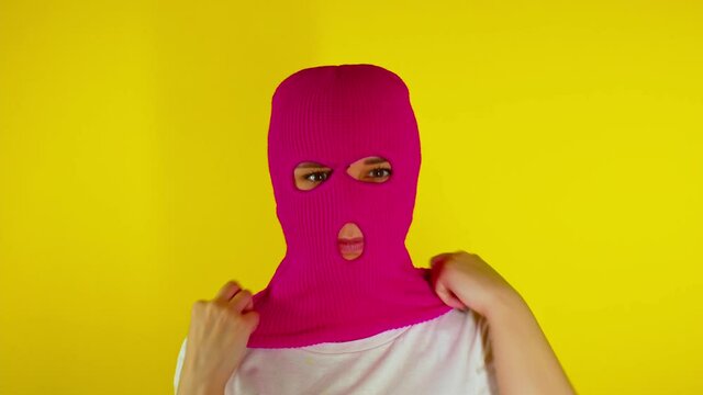Close up young woman puts on pink balaclava on yellow background. Secretive female puts on mask, looking at camera.