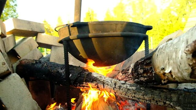 Campfire cooking. Large cast-iron cauldron on a stand. The flame from the fire heats the cauldron. Red flames. A special place for making a fire, a bonfire. Fencing made of boulders and refractory bri