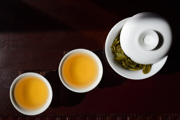 Still life and on top shot of two Chinese tea cups with golden tea and a teapot with fresh green tea leaves and a lid, on a brown table