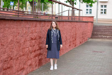 A woman in a navy blue striped dress, jacket and white sneakers stands on the pavement against a red stone wall against the backdrop of a city street.