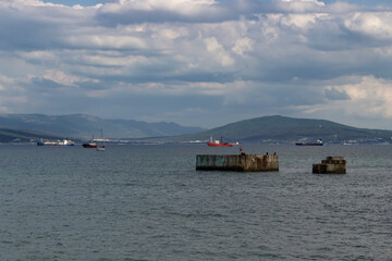 A man is fishing on the wreckage of an old pier. Ships at the entrance to the bay against the backdrop of mountains and clouds.