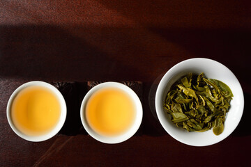 Still life and on top shot of two Chinese tea cups with golden tea and a teapot with fresh green tea leaves, side by side on a brown table