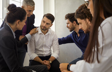Group of different people sitting in a circle supporting and comforting a sad upset young man. Concept of psychological help, group therapy and overcoming problems together