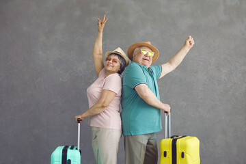 Portrait of smiling married senior couple with suitcases standing back to back on gray studio...