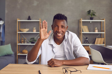 Happy black man at his desk at home saying hello and waving hand at camera. Web cam laptop computer...