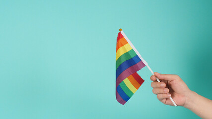 Hand is holding a rainbow flag on mint green or tiffany blue background.