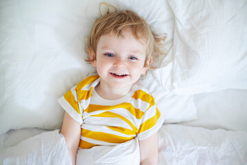 Fototapeta premium Top view of cheerful little boy in white bed early in the morning. Portrait of a handsome boy awakened