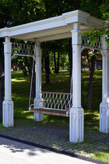 In the summer park, surrounded by greenery, there is a swing bench in the form of a gazebo with...