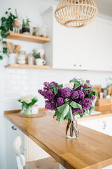 Lilac flowers in vase standing on wooden countertop in the kitchen. Modern white u-shaped kitchen in scandinavian style. Open shelves in the kitchen with plants and jars.