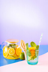 Lemonade drink of water, lemon, orange and mint leaves on multicolored background. Lime mint iced tea in a transparent teapot. Creative composition
