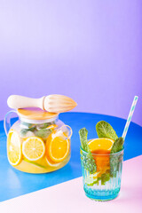 Lemonade drink of water, lemon, orange and mint leaves on multicolored background. Lime mint iced tea in a transparent teapot. Creative composition