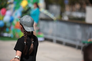 A little girl with long hair and braids in an inverted baseball cap in a black T-shirt at a sports...