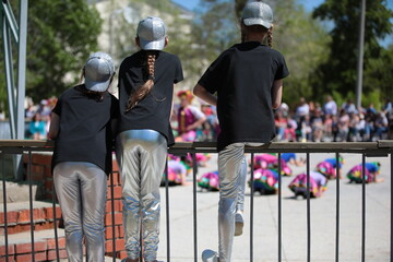 Three middle school girls in the same uniform black T shirt and silver baseball cap and leggings with long hair with a pigtail watch the performance at a children's party sitting on a fence outdoors