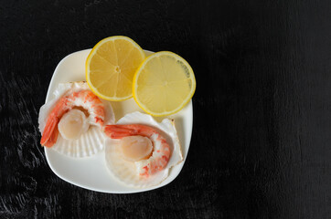 Boiled shrimp tails in a ceramic bowl with a raw scallop in the shell and lemon on a dark wooden background.