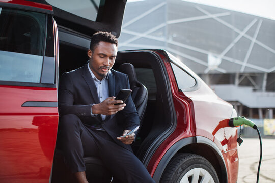 Serious African Businessman Sitting In Red Car With Opened Door And Holding Smartphone With Money Cash. Luxury Auto Is Charging. Concept Of Savings.