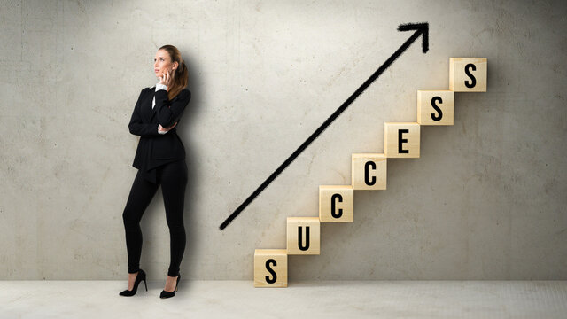 Success in business or corporate ambition concept with a stylish female businesswoman standing alongside a staircase of wooden cubes spelling SUCCESS against a concrete wall